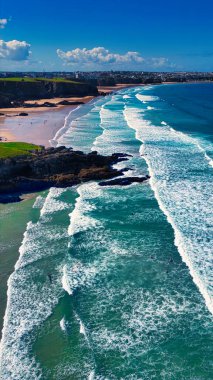 Kıyıya vuran dalgaların olduğu güzel bir kıyı şeridinin havadan görüntüsü. Plaj bazı kayalık oluşumlarla kumlu ve suda birkaç sörfçü var. Gökyüzü parlak mavi, kabarık beyaz bulutlar resim gibi bir manzara yaratıyor..