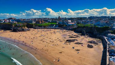 Güneşin tadını çıkaran insanlarla dolu kumlu bir sahil manzarası. Sahilde kayalık oluşumlar ve kıyıya vuran hafif dalgalar yer alıyor. Arka planda, kabarık bulutlu parlak mavi gökyüzünün altında binalar ve yeşil alanlar var..