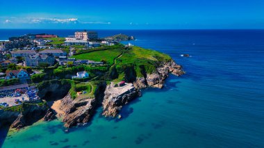 Uçurumların, kumlu plajların ve berrak mavi okyanusların bulunduğu bir kıyı manzarası. Sahne yeşil tepeler, konut binaları ve arka planda birkaç bulutlu açık mavi bir gökyüzünün altında bir otel içeriyor..