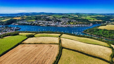 Etrafı yemyeşil tepeler ve tarlalarla çevrili pitoresk bir sahil kasabasının havadan görünüşü. Kasaba nehir kıyısında bir nehir kıyısında, benekli teknelerle kaplı. Manzarada canlı alanlar ve berrak mavi bir gökyüzü var. Huzurlu ve manzaralı bir atmosfer yaratıyor..