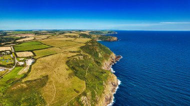 Yeşil tarlaları ve mavi okyanusa bakan kayalıkları olan engebeli bir sahil manzarası. Manzara, açık gökyüzü ve uzak tepelerle birlikte tarım arazisi ve doğal manzaranın bir karışımını içerir..