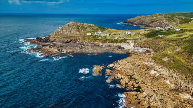 Kayalık kıyıların, berrak mavi suların ve yeşil yamaçların yer aldığı bir kıyı manzarası. Sahilde beyaz binalı küçük bir yerleşim yeri ve tepenin üzerinde bir deniz feneri görülebilir. Gökyüzü birkaç bulutla aydınlık..