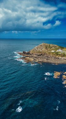 Küçük bir adası olan kayalık bir sahil şeridi. Ada, üzerinde derin mavi okyanus suları ve gökyüzünde dramatik bulutlarla çevrili bir deniz feneri barındırıyor. Sahne, doğanın ve kıyı manzaralarının güzelliğini yakalar..