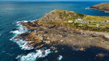 Kayalık kıyıların yer aldığı bir kıyı manzarası tepenin üzerinde bir deniz feneri ve kayalara çarpan dalgalar. Sahne, açık mavi gökyüzü ve canlı yeşil tepelerle doğanın güzelliğini yakalar..