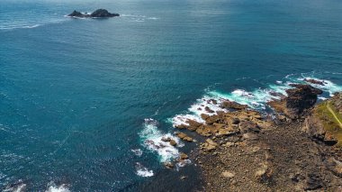 Açık mavi suları ve yumuşak dalgaları olan kayalık bir kıyı şeridi. Okyanusla çevrili ıssız bir kayalık ada uzaktan görülebilir. Kıyı şeridinde kayaların ve kumlu alanların karışımı doğal bir manzara yaratıyor..