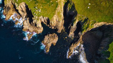 Okyanusla buluşan engebeli kıyı uçurumlarının havadan görünüşü. Manzarada kayalık oluşumlar, yemyeşil bitki örtüsü ve kıyıya vuran dalgalar yer alır. Koyu mavi su ve parlak yeşil kara arasındaki zıtlık çarpıcı bir doğa yaratıyor.