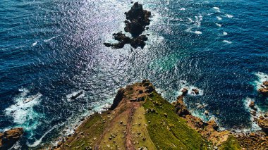 Okyanusta küçük bir adası olan kayalık bir sahil şeridinin havadan görünüşü. Su güneş ışığı altında parıldıyor, yeşil kara ve kayalık oluşumlarla güzel bir kontrast oluşturuyor. Dalgalar yavaşça kıyıya çarpar ve doğal güzelliği arttırır.