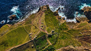Yeşil tepeleri ve kayalık çıkıntıları olan engebeli bir sahil manzarası. Okyanus dalgaları kıyıya çarpar ve kara ile deniz arasında güzel bir kontrast yaratır. Bölgenin doğal güzelliğini gözler önüne seren yollar peyzaj boyunca esiyor..
