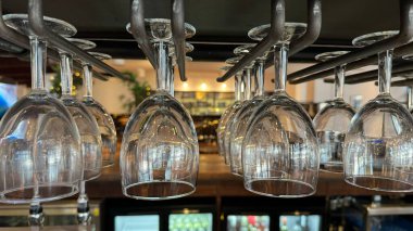 Bir barda ya da restoranda baş aşağı asılı şarap kadehlerinin yakın plan görüntüsü. Bardaklar temiz ve ışığı yansıtıyor, görsel olarak çekici bir düzenleme yaratıyor. Arka plan bulanık, canlı bir atmosfer olduğunu gösteriyor..