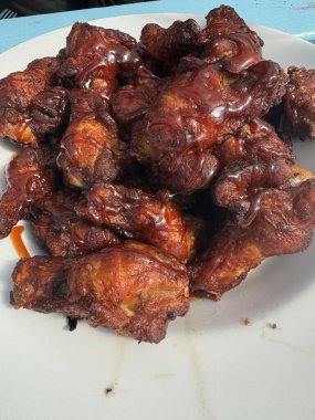 Bir tabak çıtır, ızgara kaplı tavuk kanadı üst üste yığılmış. Kanatlar koyu kahverengi ve parlak, zengin bir sosla, nefis bir şekilde yanmış bir görünüm sergiliyorlar. Tabak beyaz ve arka plan açık mavi bir yüzey..