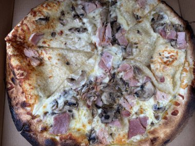 Jambon, mantar ve peynirli taze pişmiş pizza. Kenarları altın kahverengidir ve biraz kömürleşmiştir, malzemelerin bir karışımı yüzeye eşit olarak yayılmıştır. Pizza dilimlere ayrılır. Lezzetli malzemeler ortaya çıkar.