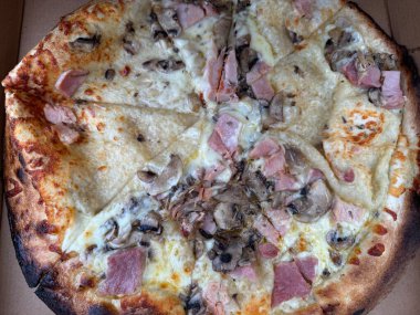 Jambon, mantar ve kremalı beyaz soslu taze pişmiş pizza. Pizza dilimlere ayrılır, altın bir kabuk ve bol miktarda malzeme sergilenir. Kenarları biraz kömürleşmiş, kırsal görünümüne ekleniyor..