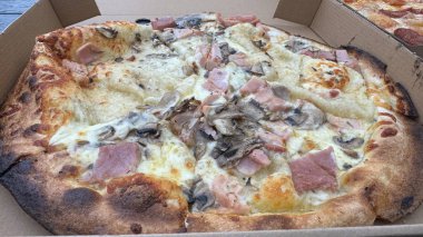 Jambon, mantar ve kremalı peynirli bir pizzanın yakın çekim görüntüsü. Kabuğu altın kahverengidir ve biraz kömürleşmiştir. Erimiş peynir ve malzemelerin karışımı görülebilir. Pizza karton bir kutuya konmuş. Paket servisi ya da paket servisi olduğunu gösteriyor.