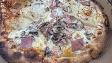 Jambon ve mantarla kaplı bir pizzanın yakın plan görüntüsü. İçinde altın-kahverengi kabuk ve erimiş peynir var. Pizza bir karton kutuya konur ve iştah açıcı görünümünü gösterir..