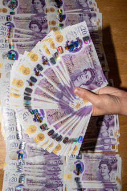 Tahta bir yüzeye daha fazla banknot serpiştirilmiş, 20 poundluk banknotlardan oluşan büyük bir yelpazeyi tutan bir el. Notlar mor ve Kraliçe II. Elizabeth 'in canlı renklerini ve holografik elementlerini gösteren bir portresi içeriyor..