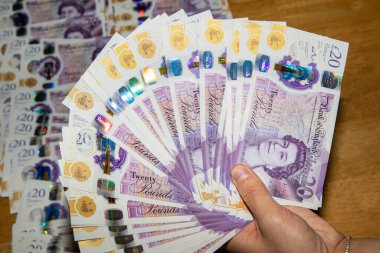 Bir el, 20 pound 'luk banknotların hayranını tutuyor, canlı renkleri ve para biriminin holografik özelliklerini sergiliyor. Arkaplanda, tahtadan bir yüzeye ek banknotlar seriliyor..