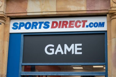 SportsDirect.com için bir tabela ve 'GAME' sözcüğünü gösteren bir pencere. Giriş mavi bir kenarlıkla çerçevelenmiş ve arka planda desenli taş bir görünüş var..