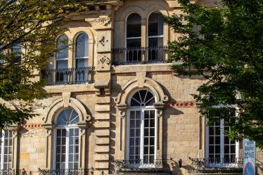 Süslü mimari detayların, dekoratif çerçeveli büyük pencerelerin ve balkonların yer aldığı tarihi bir binanın yakın çekimi. Bina, açık mavi gökyüzüne karşı klasik tasarımını vurgulayan yemyeşil ağaçlarla çevrili..