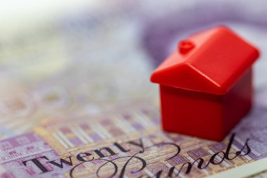 Kırmızı plastik bir oyuncak ev yirmi pound 'luk bir banknotun üzerinde duruyor, konutun maliyetini sembolize ediyor. Faturada yirmi kelimesi görünüyor..