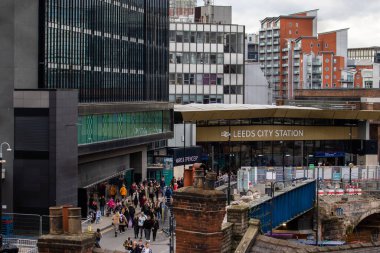 Leeds Şehir İstasyonu 'nun önünde kalabalık bir sokak sahnesi. Girişten geçen bir kalabalık var. İstasyon cephesinde istasyon adı ve Marks Spencer mağazası yazılı büyük bir tabela bulunuyor. Buil