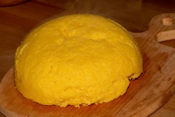 Tahta bir tahtanın üzerinde servis edilen yuvarlak, sarı bir polenta tepeciği. Doku pürüzsüz ve hafif kabarık. Buharı yeni pişirildiğini gösteriyor. Arka plan yavaşça bulanık, polentaya odaklanmış..