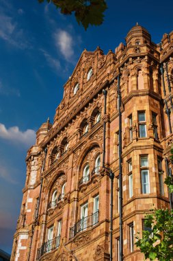 Süslü detayları olan kırmızı tuğlalı bir binanın alçak açılı görüntüsü ve işlenmiş demir parmaklıklı birden fazla pencere. Bina Manchester, İngiltere 'de beyaz bulutlu mavi bir gökyüzüne karşı kurulmuş..