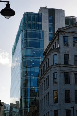 A tall modern glass building with a rounded corner stands beside a shorter stone building with ornate detailing. Both buildings are multi-story with numerous windows and are located in a city setting in Manchester, UK. clipart