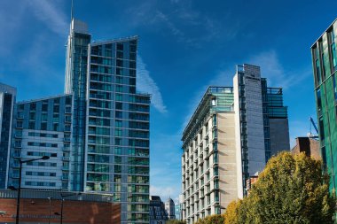 A low-angle view of three tall buildings against a bright blue sky with white clouds. The building on the left is mostly glass with some white panels, and a brown building is at its base. The building on the right is mostly stone with a glass tower a clipart