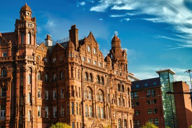 Birden fazla kulesi ve kulesi olan, beyaz bulutlu mavi gökyüzü ile çevrili, uzun ve süslü bir tuğla bina. Binanın birçok penceresi, karmaşık detayları ve Manchester, İngiltere 'de bir saat yüzü var..