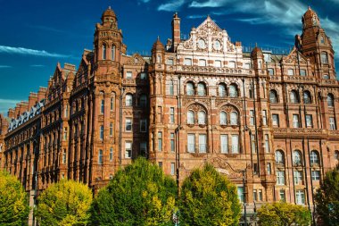 Çok sayıda kulesi ve penceresi olan, ön planda ağaçlarla kısmen gizlenmiş büyük, süslü bir tuğla bina. Gökyüzü Manchester, İngiltere 'de beyaz bulutlarla mavidir..