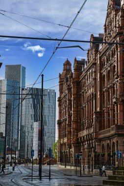 Merkezinde tramvay rayları olan, modern gökdelenler ve büyük, süslü, tuğla bir bina ile çevrili bir şehir caddesi. Gökyüzü beyaz bulutlarla mavidir ve Manchester, İngiltere 'de elektrik hatları vardır..