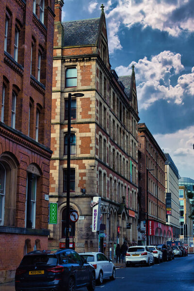 Городская улица с кирпичными зданиями, припаркованными машинами и облачным небом. Здания имеют несколько окон, а некоторые имеют вывески в Манчестере, Великобритания.