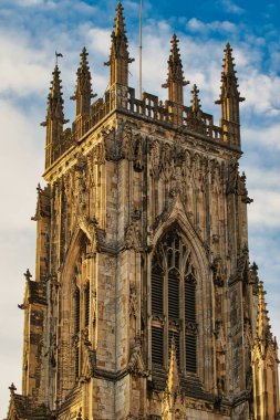 Uzun, taş bir katedral kulesinin üst kısmının yakından görüntüsü. Karmaşık gotik detaylar ve geniş kemerli bir pencere. Taş işçiliği açık ve koyu tonların bir karışımıdır ve kule York, Kuzey Yor 'da beyaz bulutlu mavi bir gökyüzü tarafından çerçevelenmiştir.