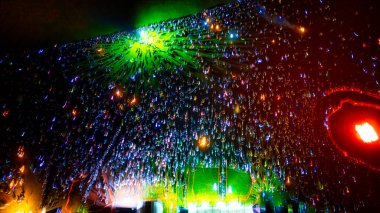 Kalabalık bir grup insan, koyu bir arkaplana karşı renkli ışık çizgileri oluşturan ışık çubukları tutuyor. Işıklar ağırlıklı olarak mavi, yeşil ve kırmızıdır. Bazı sarı ve mor ışıklar görünür..