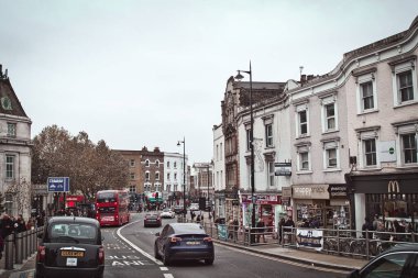 Londra 'da kırmızı çift katlı bir otobüs, siyah taksiler ve yayalar olan bir sokak manzarası. Sokaklar tuğla binalarla, bazıları da dükkanlarla dolu..