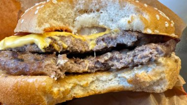 Duble çizburgerin yakın plan çekimi, kısmen yenmiş, iki köfte, erimiş peynir ve susamlı çörek gösteriliyor. Çörek hafifçe sıkıştırılmış ve hamburgerin kenarlarından peynir ve sos damlıyor..