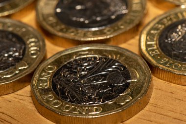 Açık kahverengi ahşap bir yüzey üzerinde birkaç İngiliz 1 poundluk madeni parasının yakın görüntüsü. Madeni paralar net bir şekilde odaklanmış, tasarım detaylarını ve metalik parıltıları sergiliyor. Alanın derinliği ön plandaki madeni parayı vurgular.