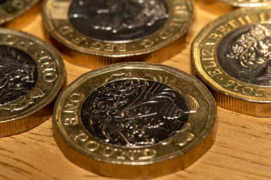 Açık kahverengi ahşap bir yüzey üzerinde birkaç İngiliz 1 poundluk madeni parasının yakın görüntüsü. Madeni paralar odak noktasında, tasarım ve yazıtların ayrıntılarını gösteriyor. Genel renk paleti altın ve koyu kahverengi / gridir.