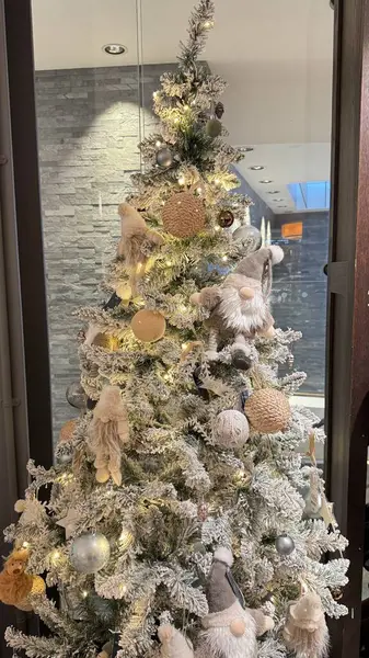 Gnomelar, yıldızlar ve küresel dekorasyonlar da dahil olmak üzere nötr tonlu süslemelerle süslenmiş bir Noel ağacı gri bir taş duvarın önünde sergileniyor. Ilık beyaz ışıklar ağacı aydınlatır..