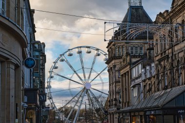 A Ferris wheel is situated between two rows of buildings on a partly cloudy day.  Overhead wires and festive lights are visible. The buildings are multi-storied and appear to be in a city center. clipart