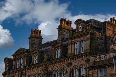 Parçalı bulutlu bir gökyüzünün altında taş bina cephesi. Binada birden fazla baca, kemerli pencereler ve süslü taş işçiliği yer alıyor. Sahne gün ışığıyla aydınlanır..