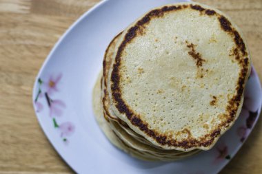 Kare üzerinde açık kahverengi bir krep yığını, çiçek desenli bir plaka tahta bir yüzey üzerinde duruyor. En üstteki krep hafif kahverengili bir çevreyi gösteriyor..