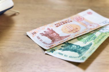 İki Moldova Leu banknotu, bir onluk ve bir yirmilik, açık kahverengi ahşap bir yüzey üzerinde uzanır. Sol üstte beyaz bir nesnenin köşesi görünür..