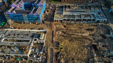 Büyük bir inşaat alanının havadan görüntüsü, yapım aşamasındaki binaları, kazılmış toprağı ve inşaat ekipmanlarını gösteriyor. Bir bina iskele ile çevrili. Kırmızı ve beyaz bariyerler görünür..