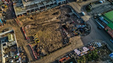 Aerial view of a construction site.  Excavated earth dominates the center, surrounded by building materials, machinery, and partially completed structures.  Roads and parking areas are visible nearby. clipart