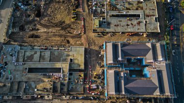 Bir inşaat sahasının havadan görünüşü iki aşamayı gösteriyor: bitmemiş beton yapılar ve neredeyse tamamlanmış koyu gri çatılı binalar, kazılmış toprak ve inşaat malzemeleriyle çevrili..