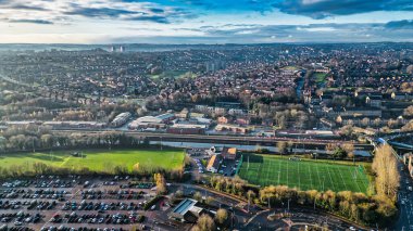 Bir kasabanın havadan görünüşü, konut alanları, endüstriyel binalar, bir kanal, spor sahaları ve büyük bir otopark. Görüntü, parçalı bulutlu bir gökyüzü altında yakalanan kentsel ve doğal elementlerin bir karışımını içeriyor..