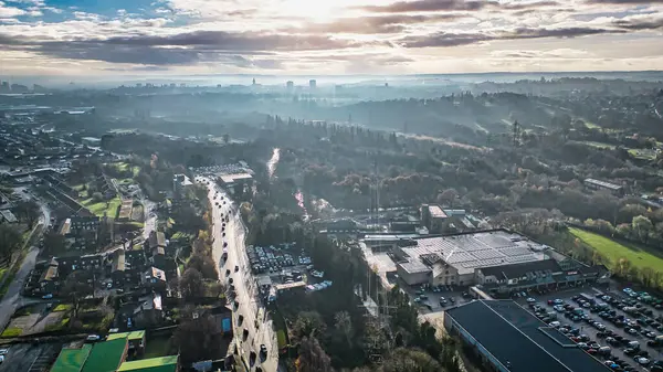 Geniş bir süpermarket, yol, konut binaları ve arka planda puslu bir şehir manzarası olan bir banliyö bölgesinin havadan görüntüsü. Görüntü, parçalı bulutlu bir gökyüzü altında kentsel ve doğal elementlerin bir karışımını gösteriyor..