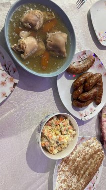 Masa düzeninde çeşitli yemekler sergilenmektedir: havuçlu jöleli et, kızarmış sosis, sebze salatası (Olivier salatası gibi) ve katmanlı kek. Masa örtüsü açık bej ve tabaklarda çiçek desenleri var..