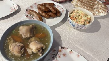 İçinde bir kase aspic, bir tabak sosis, bir kase salata ve hamur işi olan bir masa seti. Masa örtüsü açık bej renginde ve tabakta çiçek desenleri var..